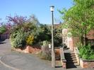 Pavement with streetlamp in foreground.
Steps between low brick walls lead to path along the front of houses.
Garage block on left behind shrubbery.
Houses on right behind small tree.
Neighbourhood Watch sign on streetlamp.