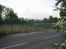 Road with high steel paling fence on the far side.
Area of scrubby gravel behind fence.