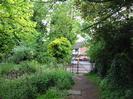 Footpath with trees on each side.
Metal kissing-gate to Mill Lane.
Houses and cars in the distance.