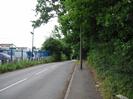 Looking south on Hitcham Road.
Bollards and blue metal fence on far side of road.
Tarmac pavement with streetlight pole.
Bushes and trees overhanging path on right.