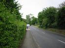 Looking west on Institute Road.
Hedge and footpath on left, with pedestrians approaching.
Car and van on Station Road.
Verge on right with trees.