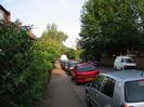 Pavement with large shrubs on the left hiding flats.
Parked cars on road.
Large trees on right.
