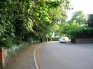 Road with tarmac pavements both sides.
Brick gatepost and low wooden slat fence on left with large trees overhanging.
Car and van parked on road with house and trees beyond.