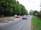 The A4 Bath Road.
Large Horse Chestnut trees on left.
Pair of traffic islands.
Cars on road.
Streetlight in right-hand verge.
Fence on right with estate-agent's signs.