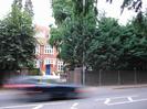 Road with passing car.
Wooden slat fence along far side of road, with trees behind the fence.
Three-storey house of red brick with white-painted window frames and bright blue door.
Street-light pole beside driveway.