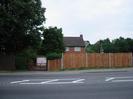 Road with trees and brown wooden slat fence on far side.
Driveway with five-bar gate leading to white garage doors.
Upper storey of house visible over the fence: red bricks, white window-frames, dark tile roof.
Roadsign warning of school.
