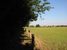 Footpath with dark trees on the left.
Barbed-wire fence with large grassy field on right.
Houses and farm buildings in the distance.