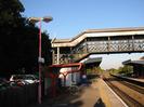 Railway platform with red covered bike stand.
Fence on left with car-park and trees behind it.
Street-light pole with Taplow sign.
Covered iron footbridge with inset GWR logos and lattice sides.
Station building with canopies beyond footbridge.