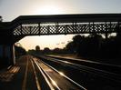 Looking west along platform 1 into the setting sun.
Iron lattice footbridge seen in silhouette.
Light glinting from main line rails.