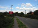 Looking north on Marsh Lane.
Dark bushes on left.
Street light with sign 'NEW TRAFFIC SIGNALS AHEAD'
Grass verge with footpath.
Roadsign warning of traffic lights.
Bushes on far side of road.