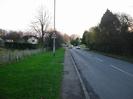 Looking south on Hitcham Road.
Wide grass verge on left with fence and houses in the distance.
Tarmac pavement.
Small trees on right.