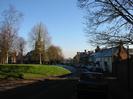 Rectory Road passing the Village Green. St Nicolas Church visible through the trees. Houses and pub in deep shadow on the right.