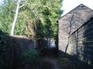 Footpath with fence and trees on the left.
Buildings with dark weatherboards on the right.
Part of High Street visible in the distance.