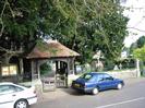 The entrance to St Nicolas Church, seen across the High Street.
Wooden gate with tile roof.
Stone walls with iron railings.
Tombstones in churchyard.
Notice board.
Parked cars.
Part of Wellbank visible in the distance on the right.