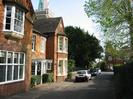 Red brick houses on left.
Part of St Nicolas Church visible over the roof.
Parked cars on High Street.
St Nicolas House visible at the bottom of the road.
Hedge around Village Green on the right.