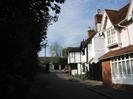 Looking North on High Street.
Dark hedge and trees on the left.
White house with part-timber walls in the centre.
Red brick house with pink upper level on the right.