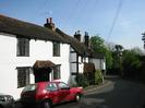 White house with black front door and tiled porch.
Cottage with white walls and black timbers.
Tip of church spire just visible between chimneys.
Dark hedge on right, with end of Village Hall roof.
Parked cars.
Telephone wires.