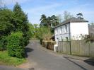 Looking East at the top of High Street.
Hedge and trees on the left.
White house with grey roof on the right.
Wooden fence.
Junction with Hill Farm Road in the distance.