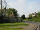 Looking North on Hill Farm Road.
Grass area with street sign for Desborough Cottages.
Wooden fence on left with house visible beyond.
Road sign for crossroads.
Bus stop, telephone pole, and houses partly hidden by hedge on the right.