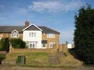 House built from sandy-coloured bricks. Partly rendered white.
Dark slate roof.
Low brick wall at top of bank beside road.
Steps from road to house.
Green telephone junction cabinet.
Dark Cypress trees on the right.