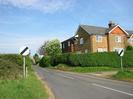 Looking East on Hitcham Lane.
Hedges both sides of road.
National speed-limit signs.
House behind hedge on right.
Trees in the distance.
Telephone wires.