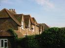 View along the rear of Church Cottages.
Sandy-coloured bricks, red clay tiles.
White-painted downpipes.
High ivy-covered fence.
Chimneypots on skyline.