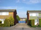 Road leading to block of garages.
Houses each side of the road.
Garages have blue doors, and more houses are visible over them.
Hedges in fornt of houses.