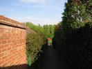 Footpath.
High brick wall and hedge on the left.
Hedge on the right.
Grass area visible beyond barriers.
Red car parked on Hill Farm Road.