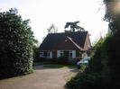 Concrete drive between high hedges leading to house.
House has large windows on the ground floor and is built of brick with tile roof.
Large trees visible over the roof.