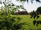 Cricket pitch seen through bushes.
Large house in the style of Lutyens on far side of pitch.