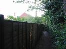 Footpath with high wooden fence on the left and bushes on the right.
Houses visible over the fence.