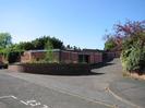 Row of garages with tarmac area in front.
Distant trees behind high brick wall on left.
Shrubs on right.
Part of a reserved parking space for disabled people visible in the foreground.