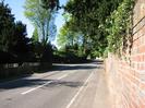 Looking north on Berry Hill.
Low fence on left side of road, with dark trees behind.
Entrance to Mill Lane, with large trees behind.
Pavement with high red-brick wall on right.