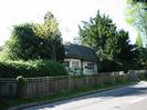 White thatched cottage.
Low wooden slat and trellis fence alongside road.
Bushes and trees around cottage.