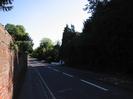 Looking south on Berry Hill.
High red-brick wall on left.
Road with traffic island in the distance.
Low white walls and gate.
Dark bank of trees on the right.