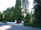 Curved white walls flanking wooden five-bar gate.
Road in foreground.
Tall trees with roof of white house just visible.