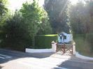 Low white walls flanking pair of gates.
Hedge and trees each side.
Small white building visible through the gates.
