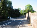 Looking north on Berry Hill.
Bank of dark trees on the left, with low white walls flanking a gate.
Pavement and high brick wall on the right.
SLOW marking on road, and start of crosshatched area for traffic island.