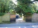 High brick walls with large black wooden gates.
Trees overhanging walls.
Gravel driveway with dark wooden buildings.
Road in foreground.