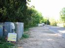 Looking north on Berry Hill.
Electricity sub-station on left with chainlink fence and stacked roadsigns.
Gravel area beside road with trees on the left.
Traffic-calming `gate features' each side of the road.