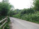 Looking west on Mill Lane.
Post and rail fence on the left with gate to public footpath.
Road with banks one each side.