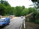 Looking east on Mill Lane.
Fence and bridge parapet on the left.
Steel safety barrier and old overgrown building on the right.
Large trees in the background.