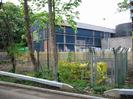 Paper mill buildings: large blue metal-clad building with pipework and tanks.
Chainlink fence around yellow pipework in the foreground.