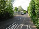 Looking north on Mill Lane.
High metal fence on the left with trees beyond it.
Trees and bushes on the right.
House visible in the distance.