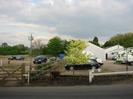 Car-park with low railings and trees beyond.
Low metal-clad buildings on right.