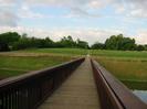 Wide wooden footbridge across river.
Field on far side rises towards a bank of trees.