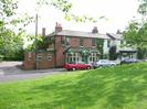 Oak and Saw public house.
Red brick building with slate roof.
Pub sign with golden cockerel on top.
Pub car park on the left.
White houses to the right.
Telephone pole set in pavement by pub.
Parked cars.
Part of Village Green in the foreground.