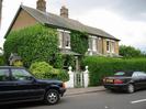 Group of houses with parked cars on road in front.
Houses are built of yellowish brick, and have dark slate roofs.
White wrought-iron gate between white pillars, each of which has a round decorative top-piece.