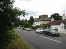 Road with grass verge and trees on left.
Houses and parked cars on right.