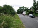 Road with grass verge and trees on left.
Telephone pole, roadsign, and postbox.
Houses and trees on the right.
Cars on road.